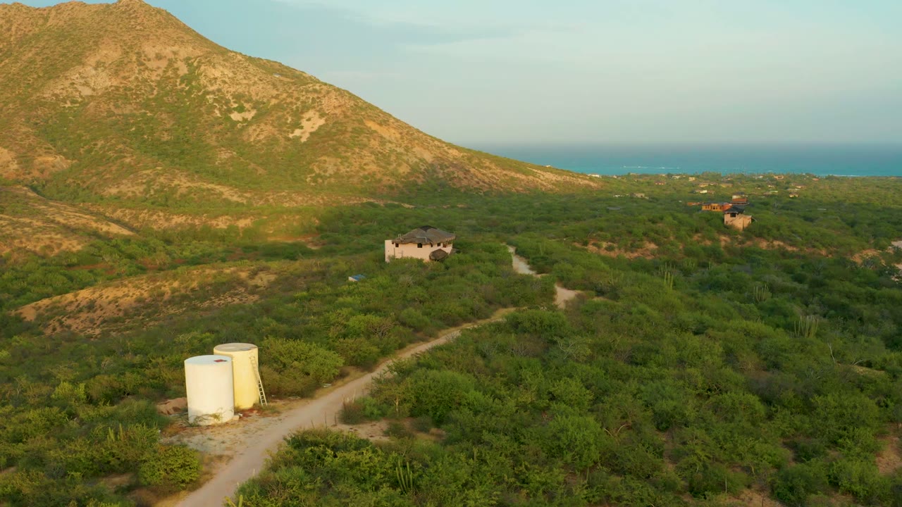 Opening frame over Cabo Pulmo, Baja California Sur, showing a dirt road through green vegetation, two white tanks in the lower left, a small building near the mountain slope, distant sea, and no visible people.
