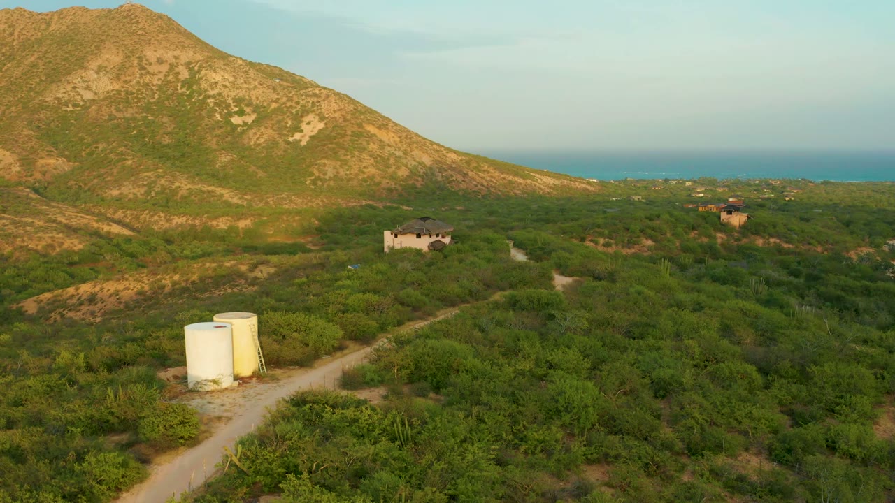 Mid-clip frame over Cabo Pulmo, Baja California Sur, where the dirt road angles past the white tanks, the small building sits left of center, the mountain fills the upper left, the sea runs across the horizon, and no visible people appear.