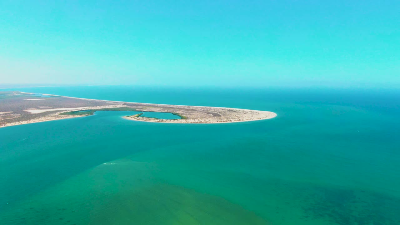At 0:00, a high aerial view over San Carlos, Baja California Sur shows a narrow sand island near the horizon, turquoise water in the foreground, and no visible people.