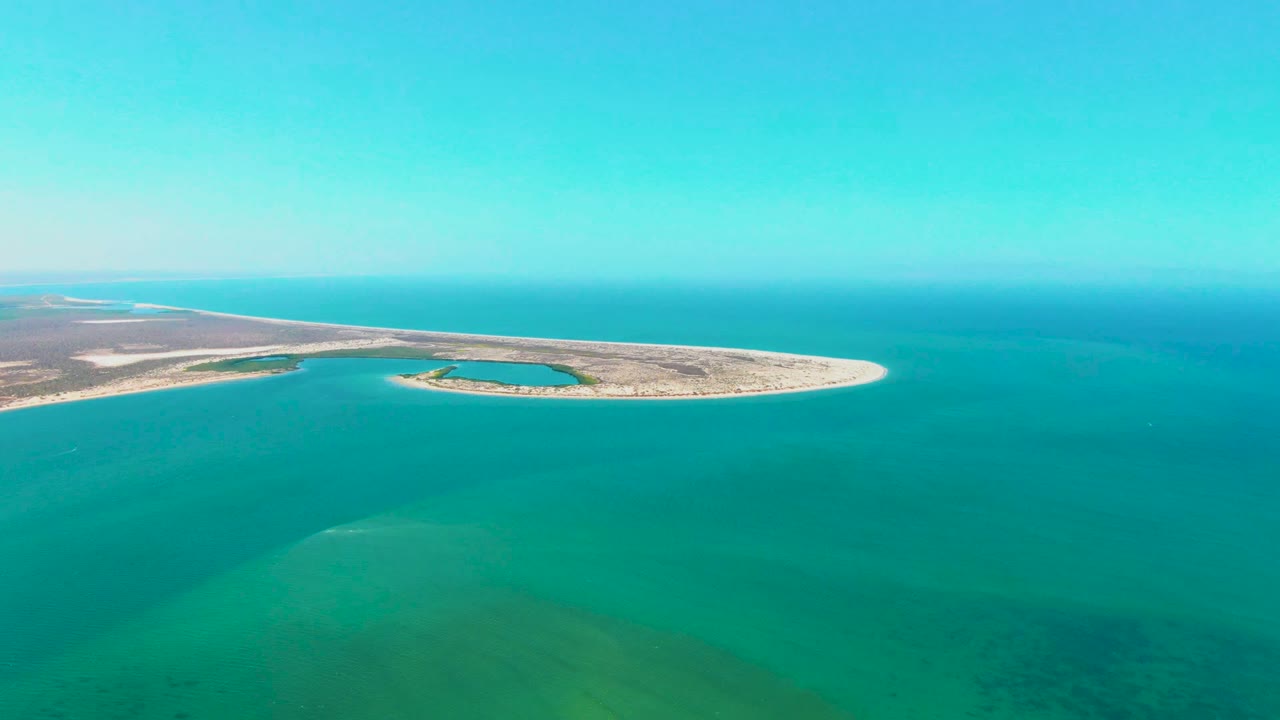 At 0:01, the aerial frame keeps the San Carlos sand island across the upper image with a small blue-green water pocket inside the shoreline and no visible people.