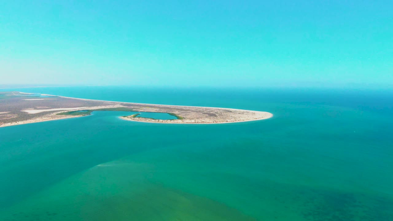 At 0:02, the drone view over San Carlos holds a wide composition with the curved island extending right, layered blue-green water below, and no visible people.