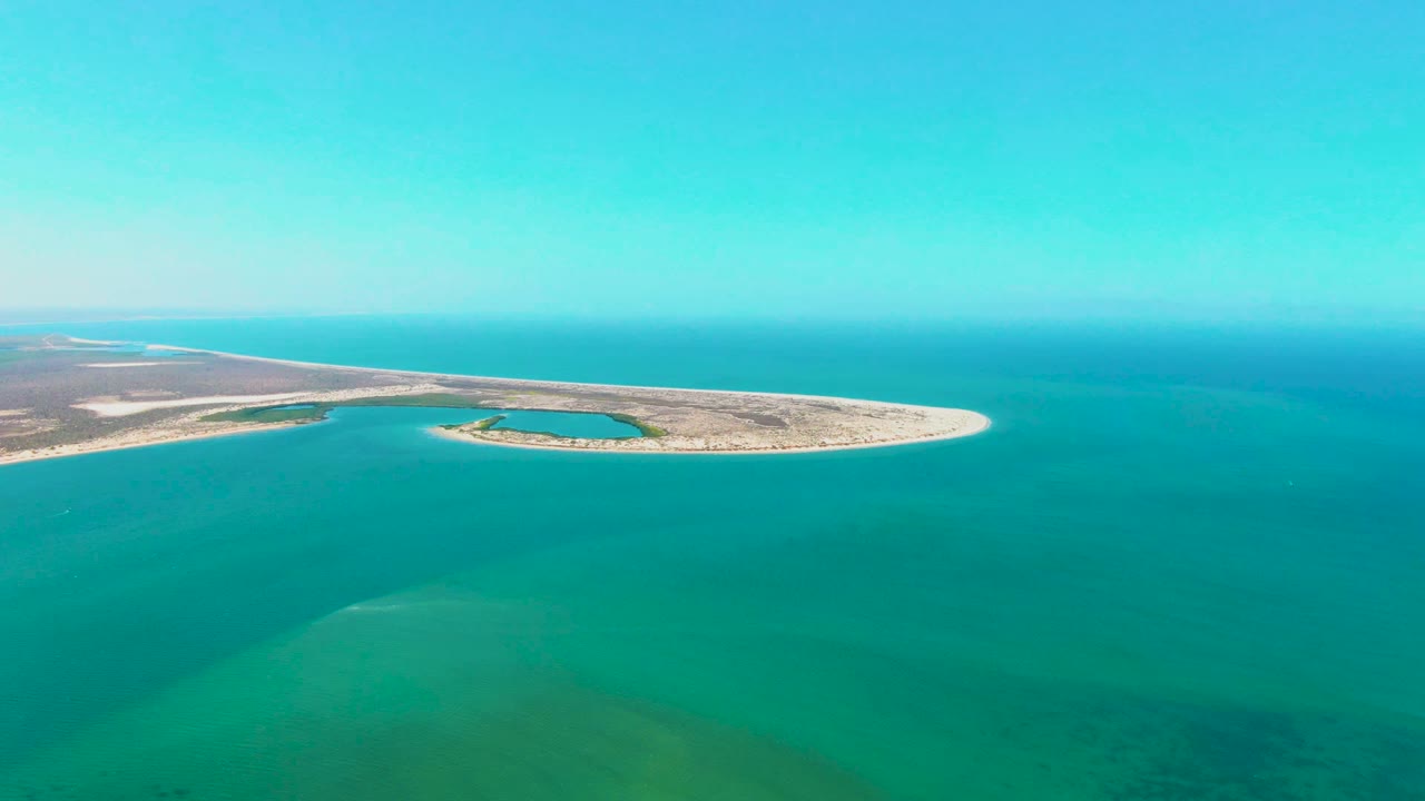 Aerial coastal view with a low sand island and turquoise shallows in the foreground.