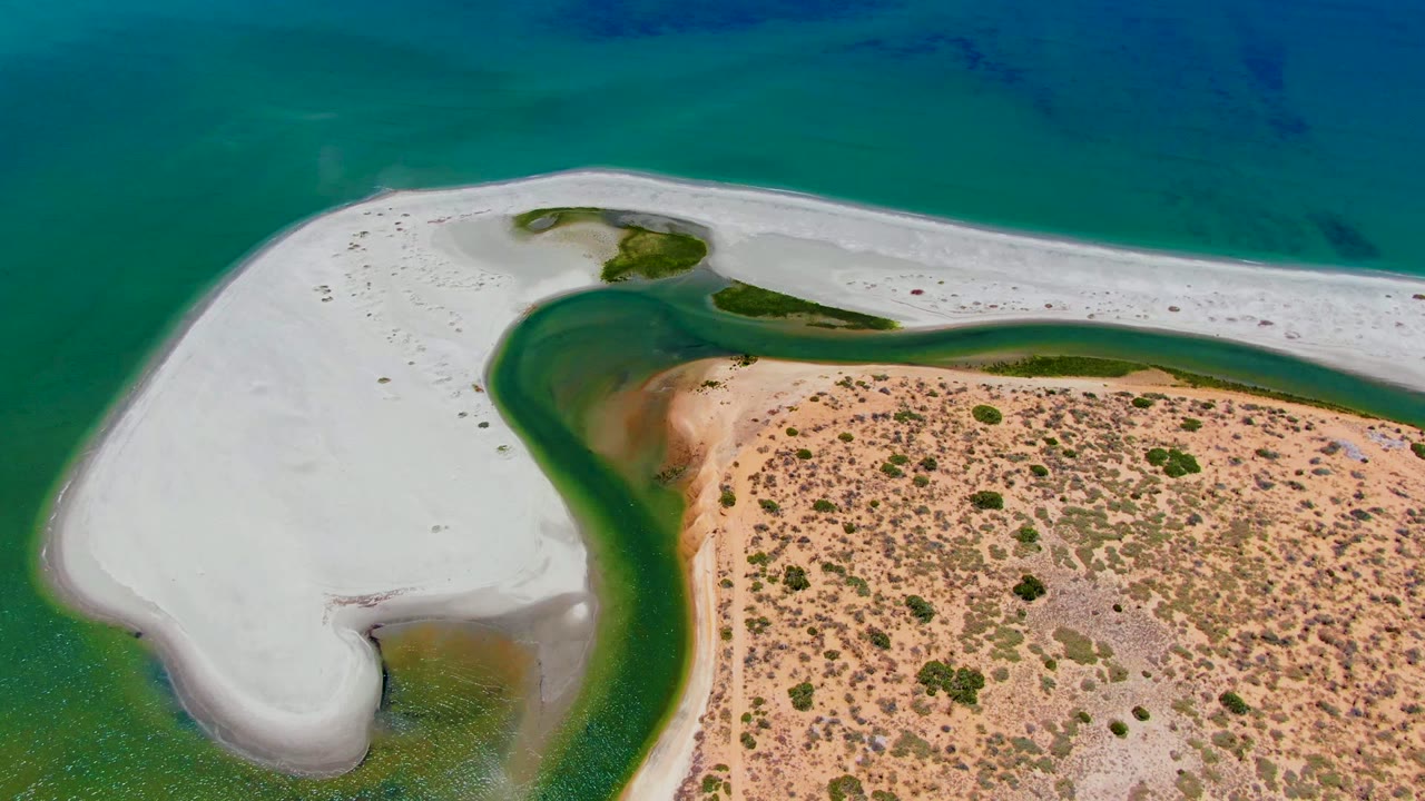 At two seconds, the San Carlos coastline remains framed from above with bright white sand, a darker green channel, tan inland sand, scattered low vegetation, and no visible people.