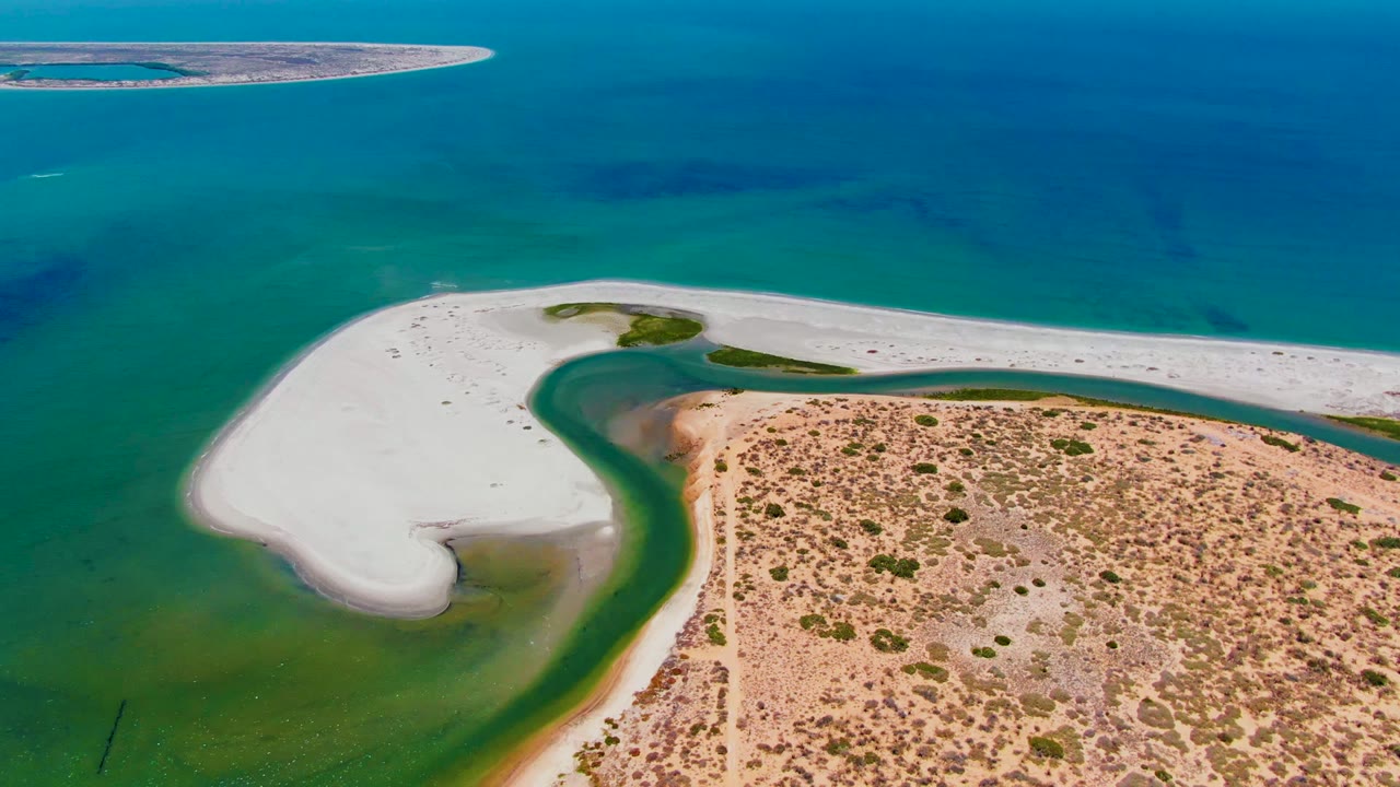 Aerial frame over San Carlos, Baja California Sur, with the curved white sandbar more centered around the green lagoon and dry vegetated land below, with no visible people.