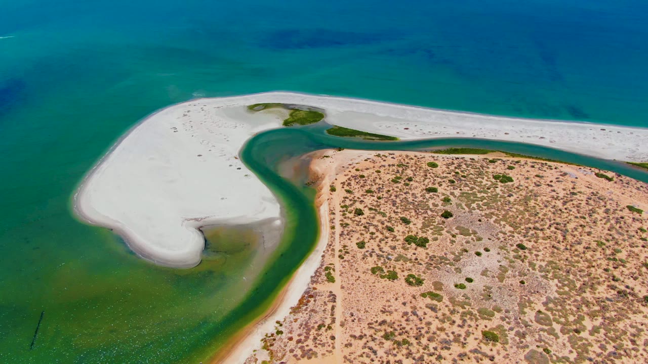 Overhead shoreline view with a white sand hook, green lagoon water, and desert scrub.