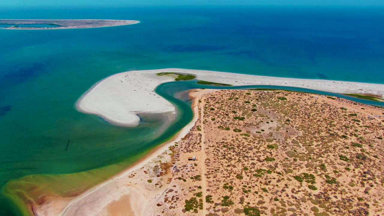 Aerial frame over San Carlos, Baja California Sur showing a curved white sandbar beside green shallow water, scrubby sandy shore, a dirt road, distant blue ocean, and no visible people.