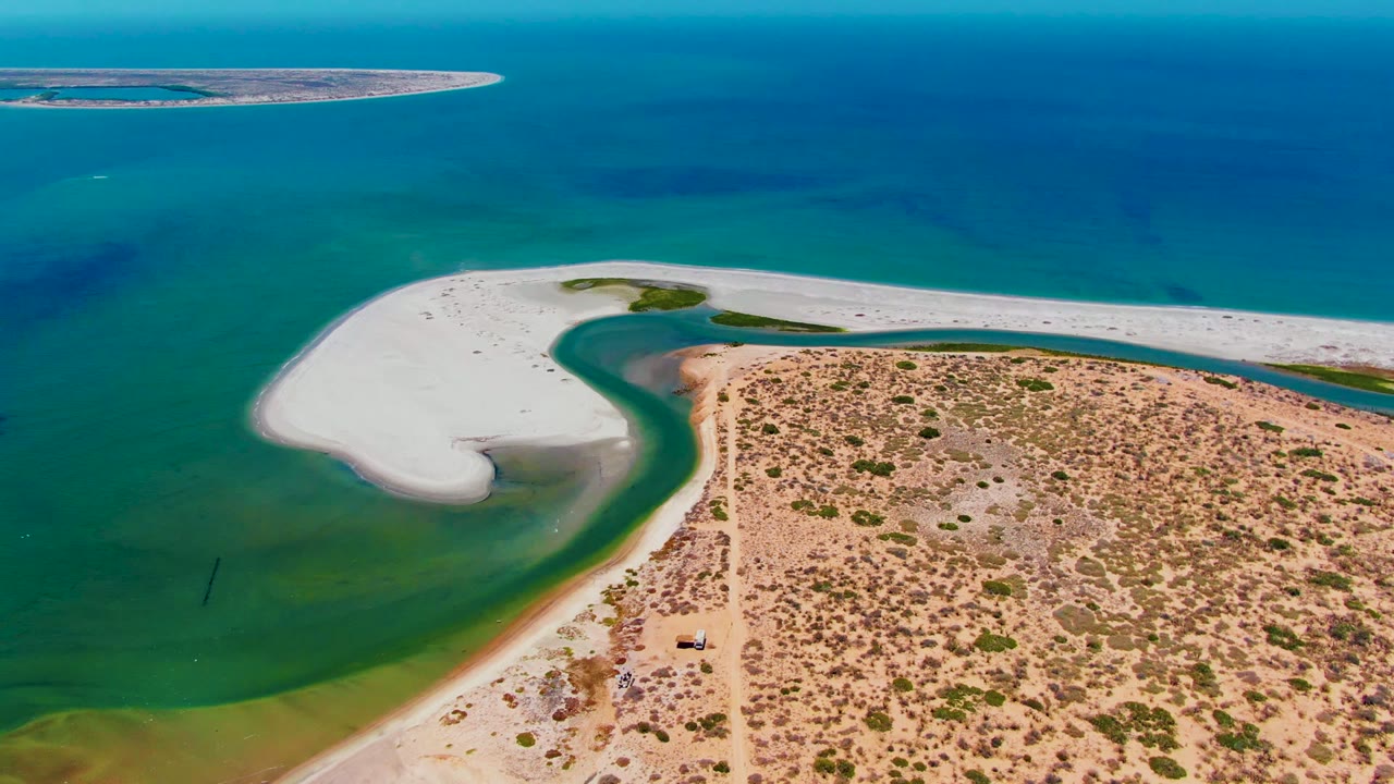San Carlos, Baja California Sur aerial frame showing the white sandbar sweeping across the middle, green and blue water gradients, scrub-covered sandy land, a dirt track, and no visible people.