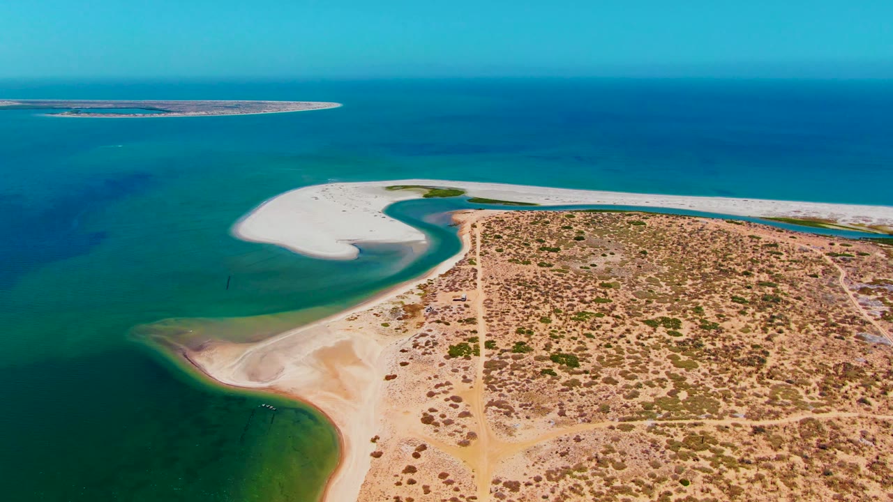 Opening aerial frame over San Carlos, Baja California Sur, showing a curved white sandbar between turquoise shallows and dry scrubland, with a distant island near the horizon and no visible people.