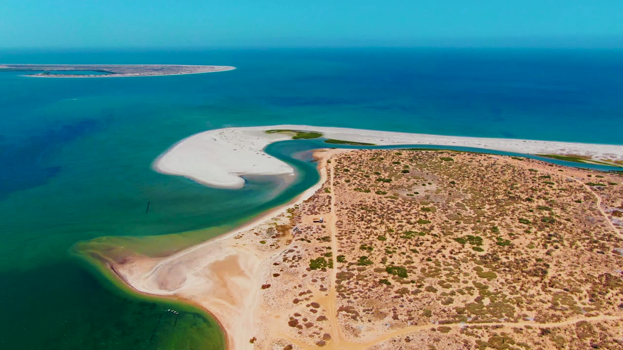 At 0:02, the aerial composition shows the white sand spit extending across the upper shoreline, a darker green shallow inlet on the left, dry Baja California Sur terrain on the right, and no visible people.