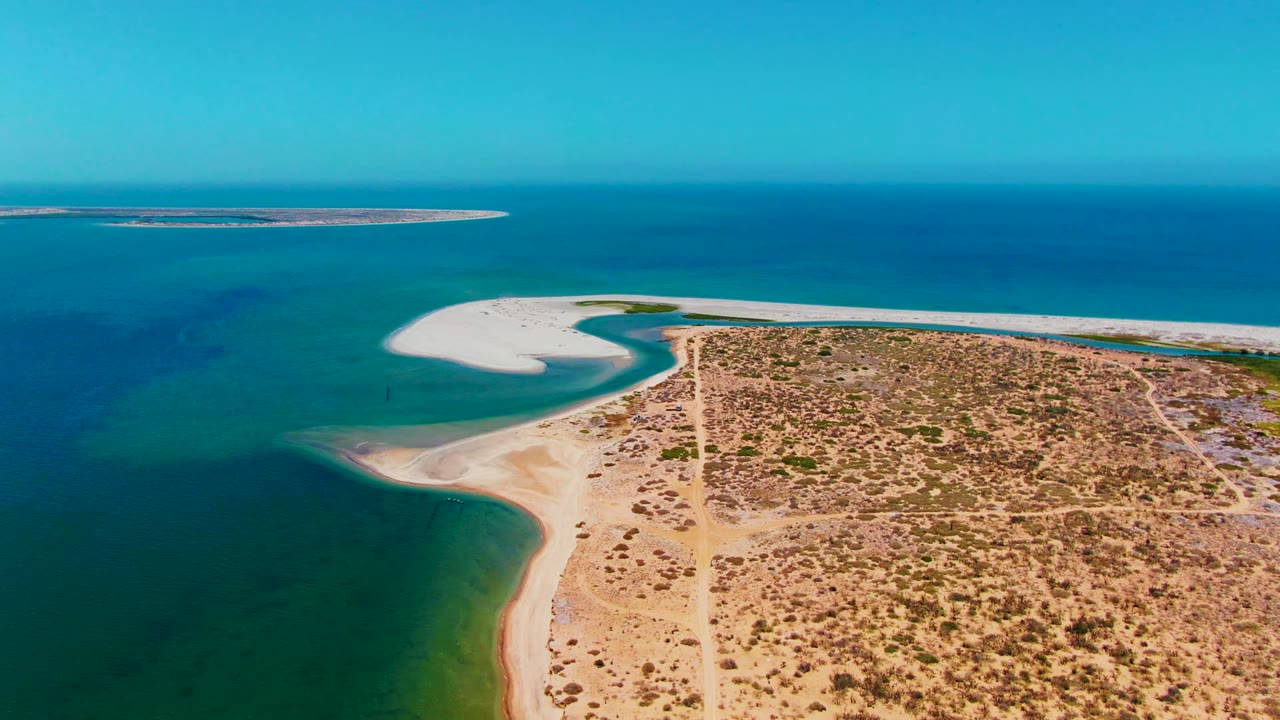 Aerial view in San Carlos, Baja California Sur, showing a curved sandy shoreline beside turquoise shallows, dry scrub terrain, and a distant offshore sandbar with no visible people.