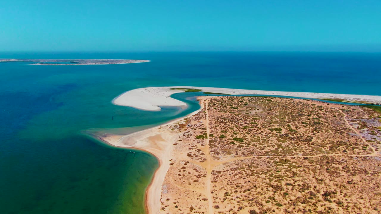 Drone view of sandy coast, green-blue water, scrubby dry land, and offshore sandbar.