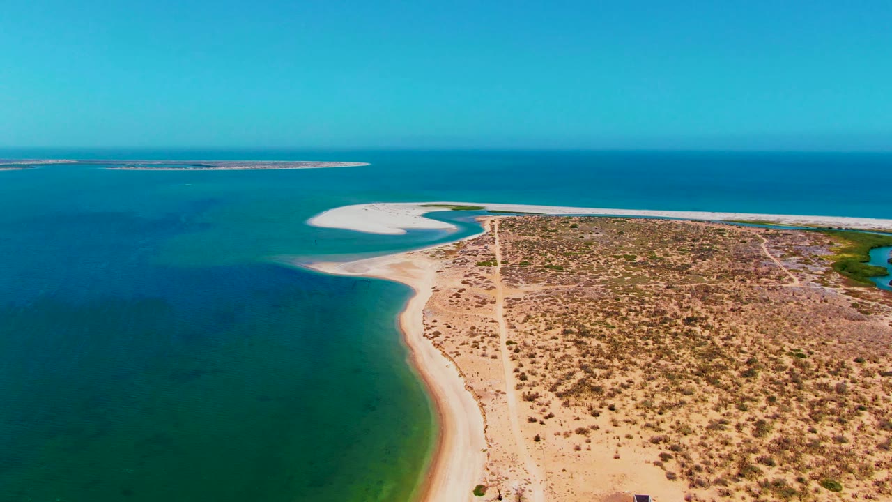 Aerial frame over San Carlos, Baja California Sur, showing blue-green coastal water, a pale sand curve, sparse scrubland, a straight dirt road, distant low land on the horizon, and no visible people.