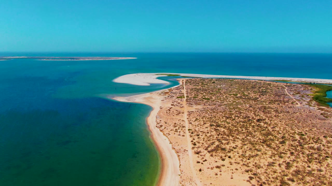 Drone view of a sandy point bordered by turquoise water and dry scrubland.