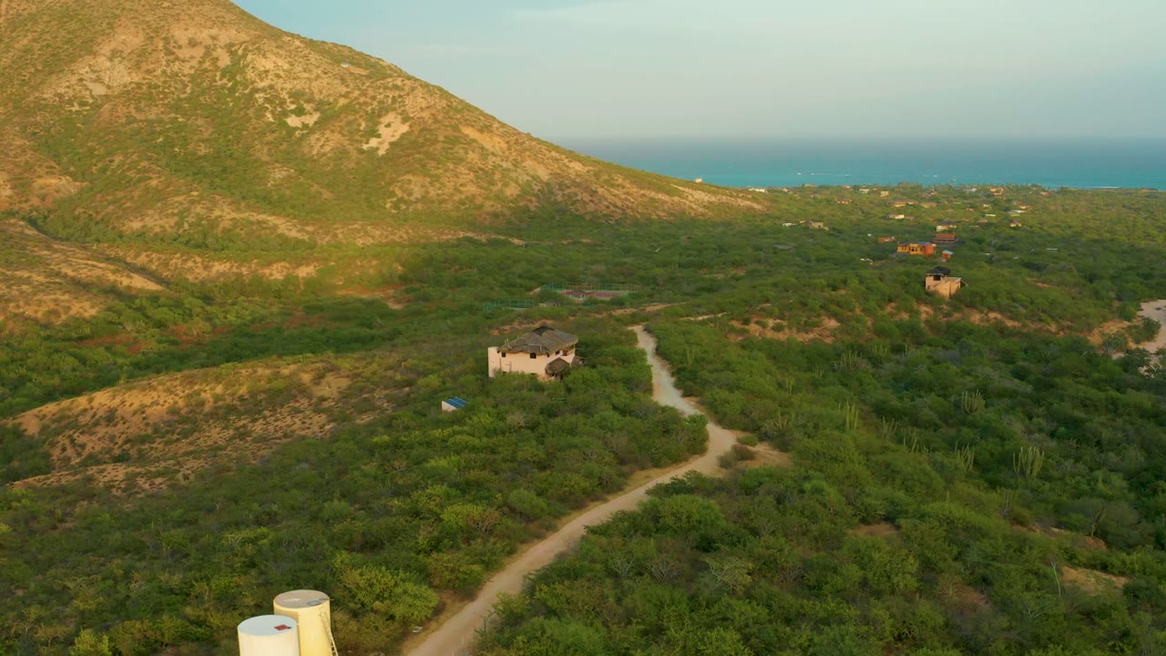 Opening aerial frame over Cabo Pulmo, Baja California Sur with two pale cylindrical tanks near a dirt road, green brush, a small house, a tan mountain slope, distant ocean, and no visible people.