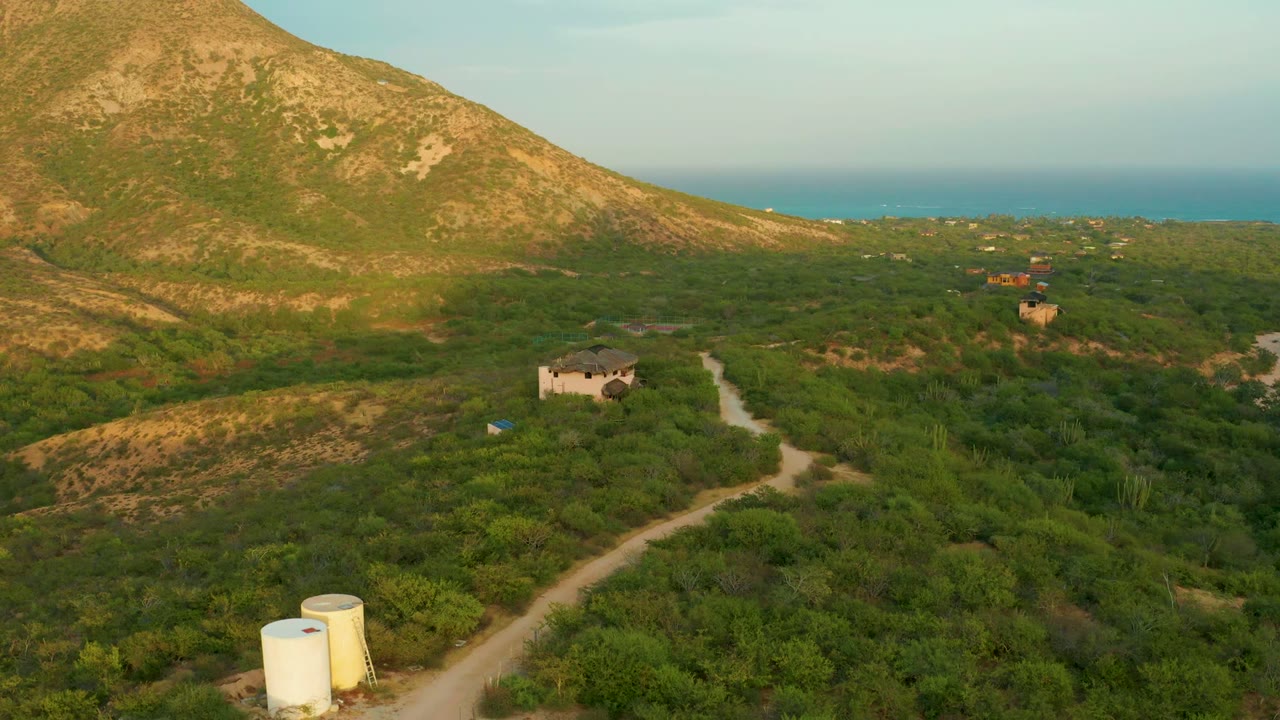 Aerial Cabo Pulmo frame with two tanks at the lower edge, a pale road cutting through green brush, small buildings in the middle distance, a mountain shoulder to the left, ocean beyond, and no visible people.
