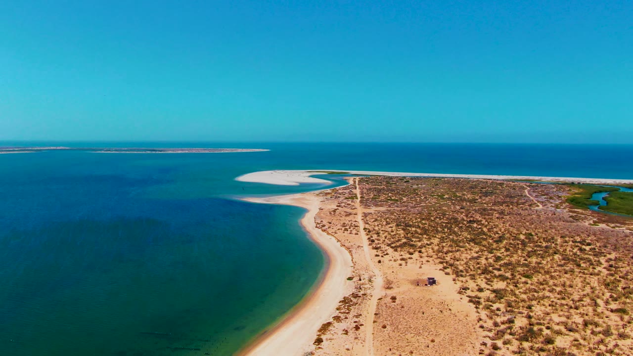 Aerial frame over San Carlos, Baja California Sur showing a sandy road running beside a curved beach, turquoise nearshore water, arid scrubland, distant blue water, and no visible people.