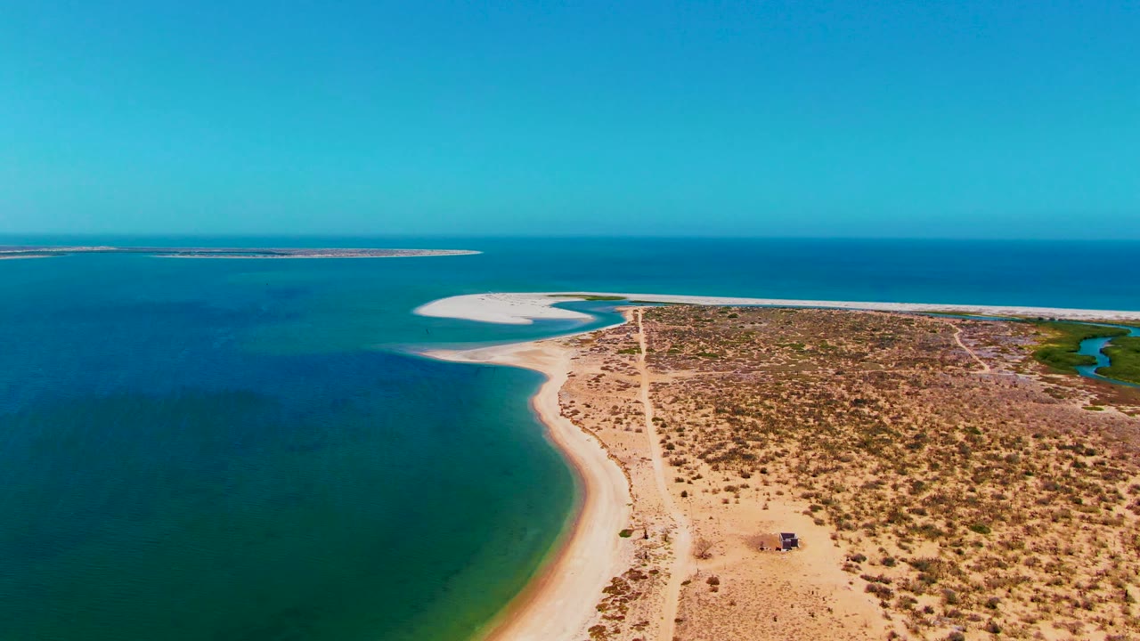 Drone-like aerial frame over San Carlos, Baja California Sur showing the curved sandy beach on the left, a straight dirt road through the land, sparse vegetation, deep blue water, and no visible people.