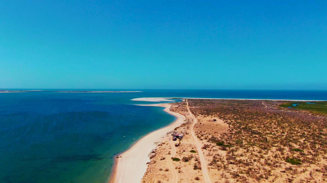 Aerial view in San Carlos, Baja California Sur, showing a pale dirt road between white sand beach and scrubland, blue ocean to the left, a small vehicle on the road, and no visible people.