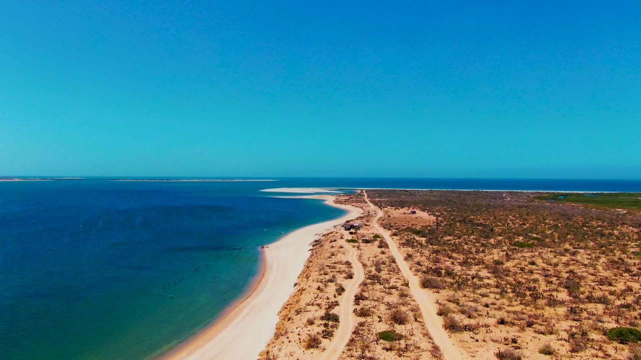 Aerial frame from San Carlos, Baja California Sur with turquoise shallows near the beach, white sand, brown scrubland, sandy tracks, and no visible people.