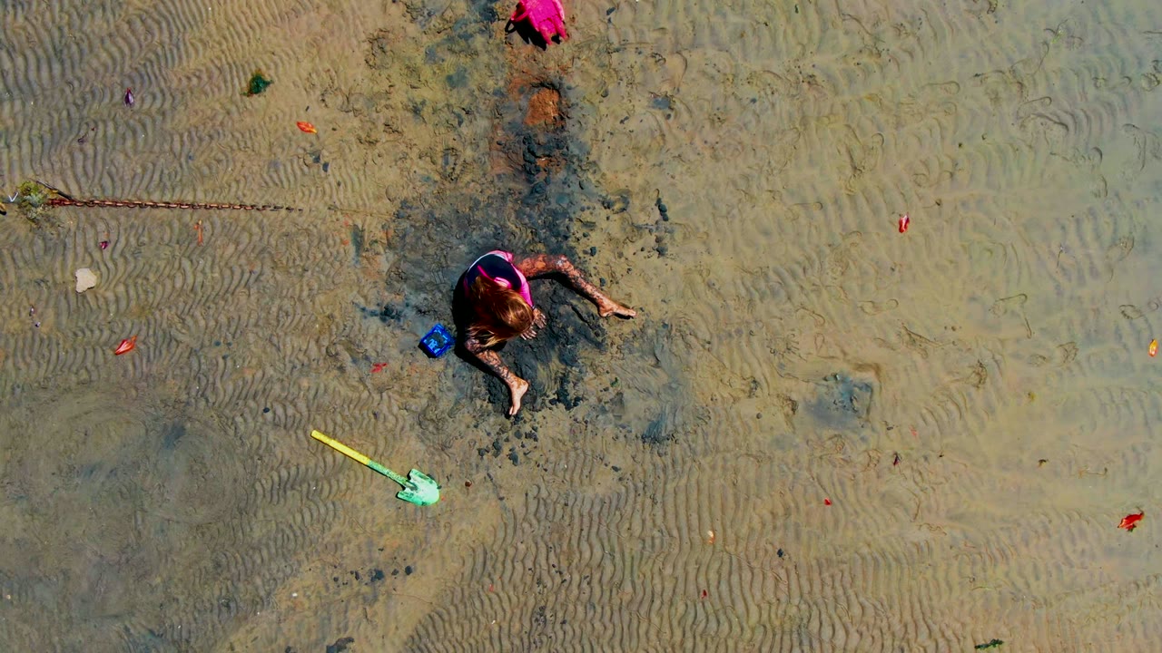 Overhead frame from San Carlos, Mexico, with a child in pink sitting on wet rippled sand near a yellow shovel, blue bucket, and dark digging patch.