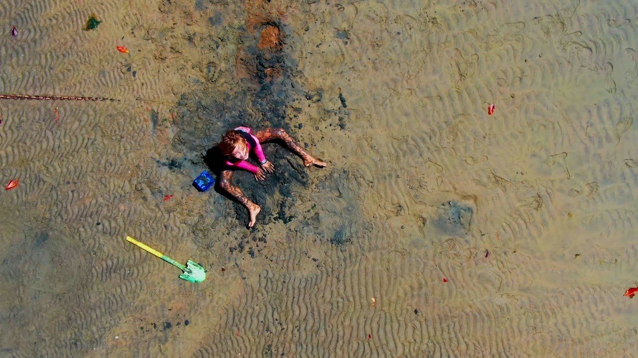 Child reclined on wet sand beside a blue bucket and yellow shovel.