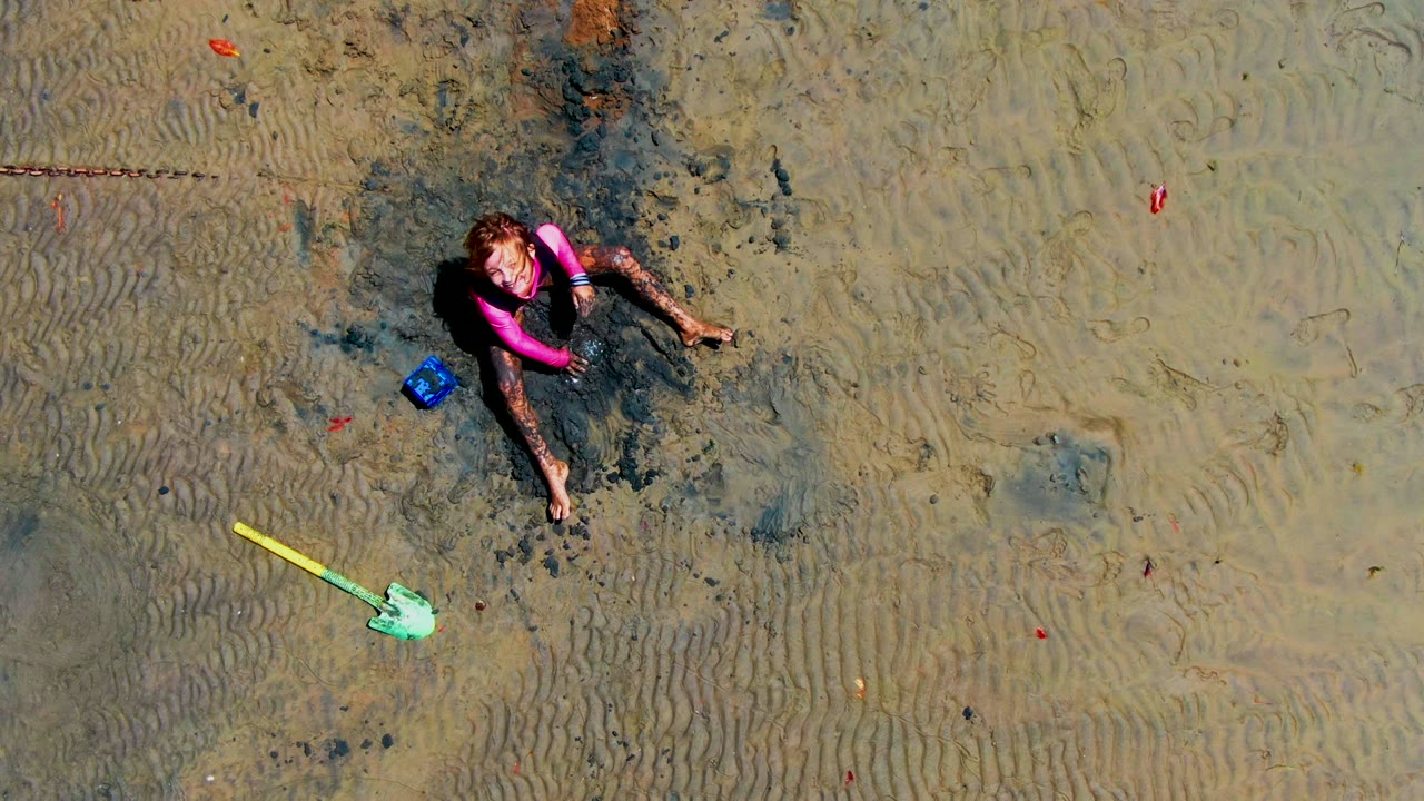 Overhead San Carlos, Mexico frame with the child turned beside a pink toy, a blue bucket, and a yellow shovel on patterned wet sand.
