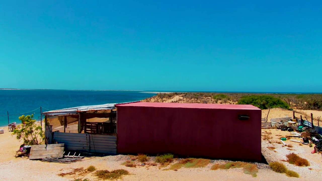 Red container structure and simple shelter on a sandy San Carlos, Baja California Sur beach with blue ocean behind them and no visible people.