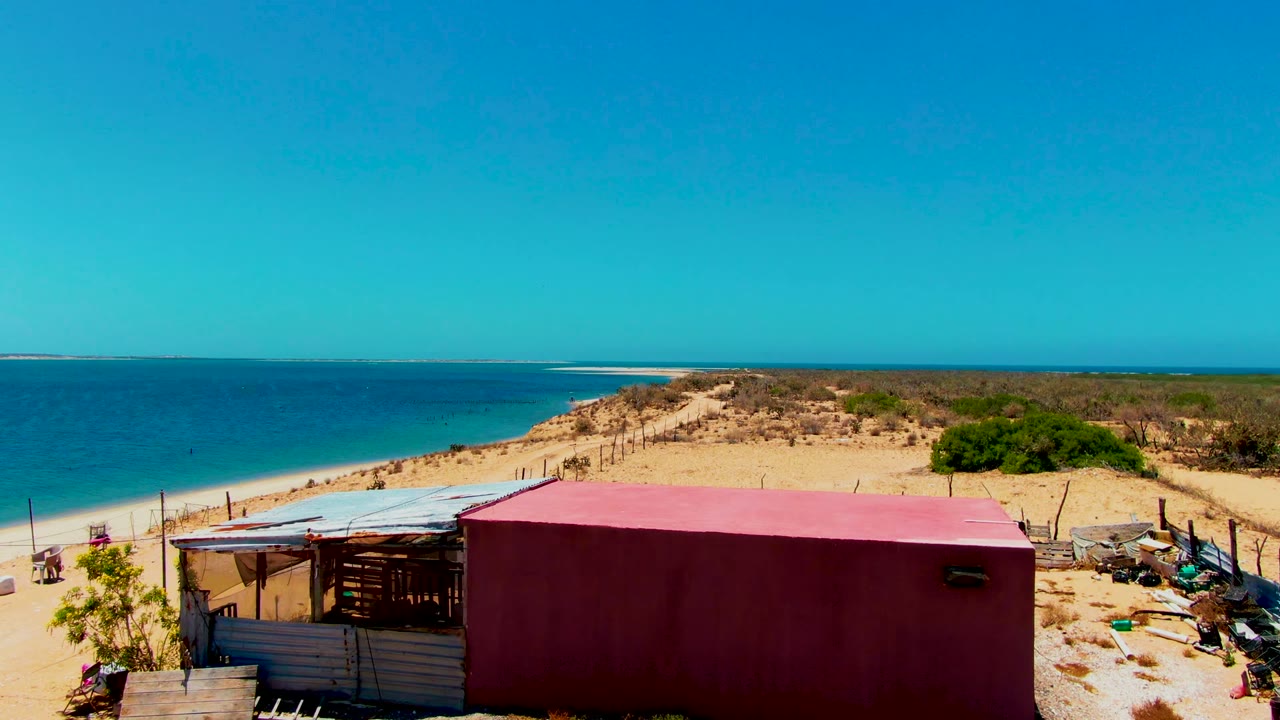 The drone view places the red structure lower in frame, with turquoise water, white sand, dunes, scrub, and a clear sky in San Carlos, Baja California Sur, with no visible people.