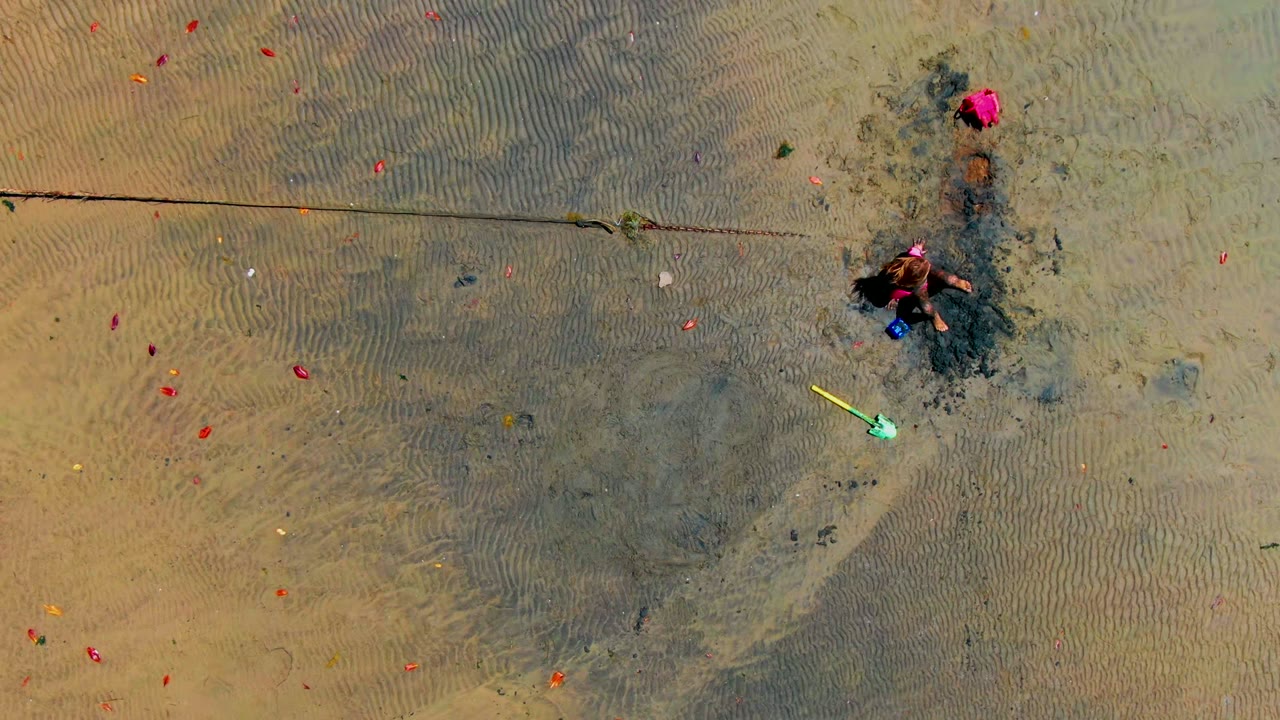 At 0:00, an overhead frame in San Carlos, Baja California Sur shows a child beside a dark wet sand patch, with a yellow shovel below and scattered red objects across the rippled beach surface.