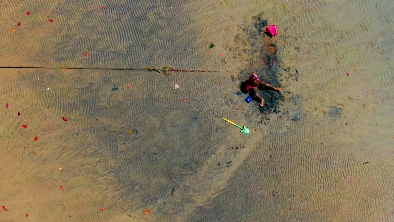 Child reaches across wet sand with a yellow shovel nearby.