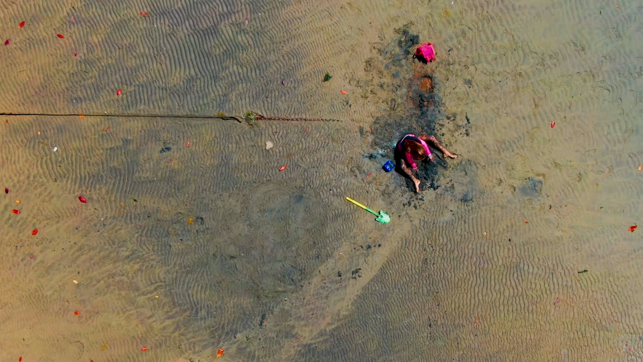 At 0:02, the child is crouched closer to the darker sand area, with the yellow shovel still visible below and small red objects dotting the shallow wet beach in San Carlos.