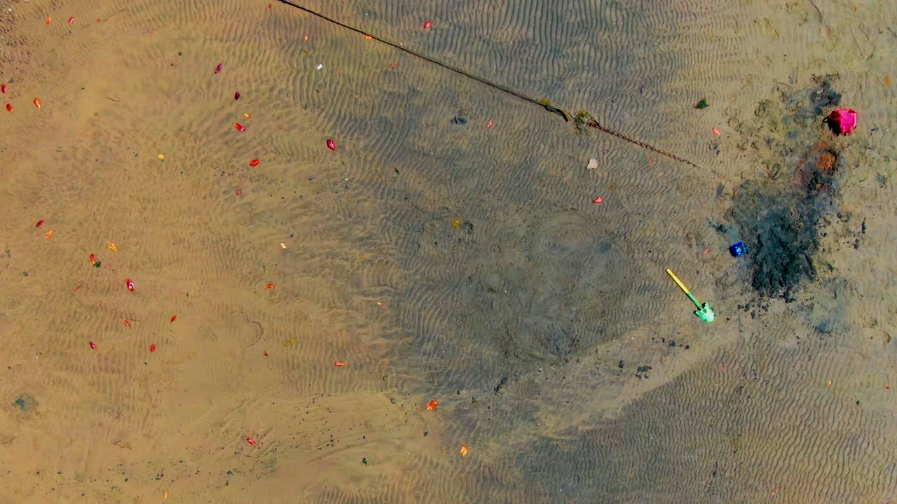 Top-down view of wet rippled sand in San Carlos, Baja California Sur, with small scattered red and orange objects, a yellow object near shallow water, and no visible people.
