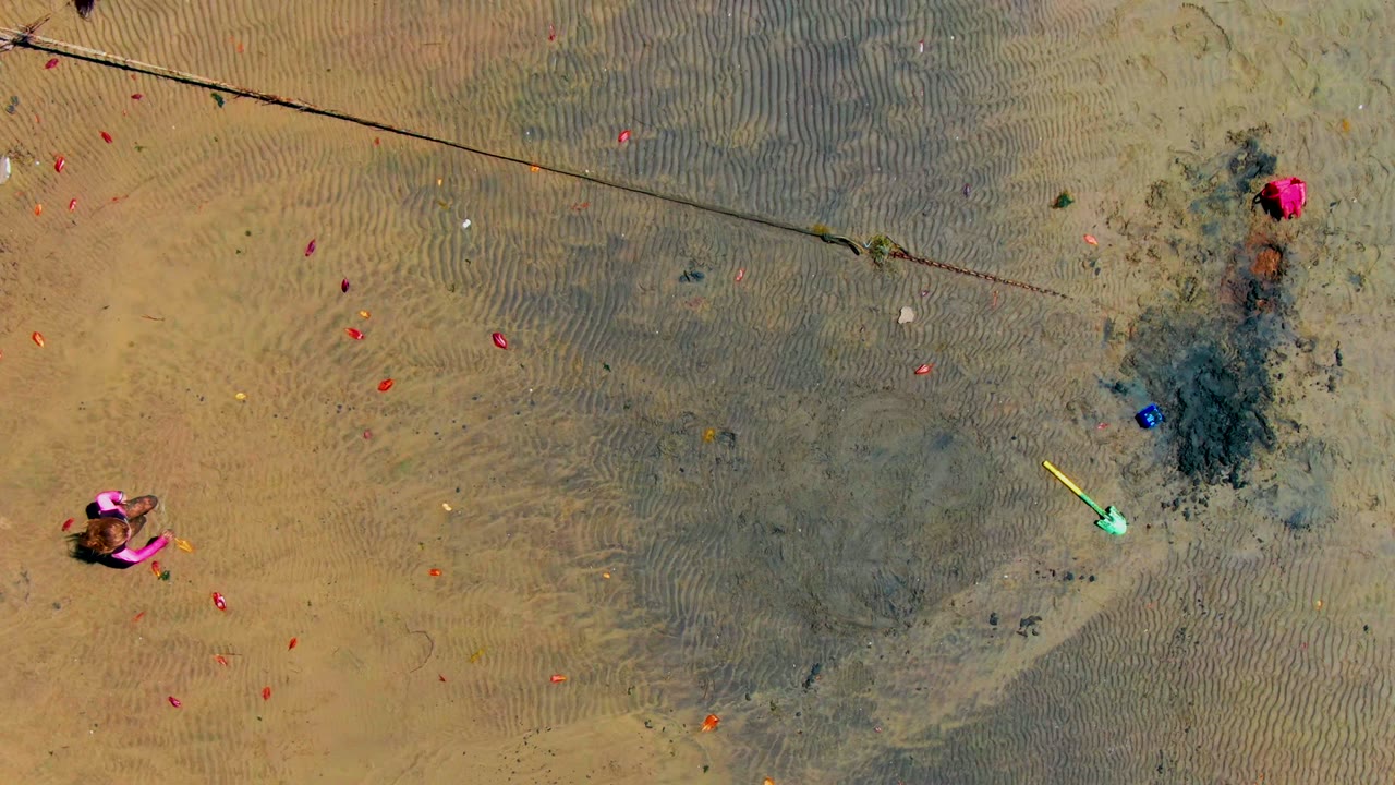 Overhead view in San Carlos showing a person in a pink top and dark pants entering the open wet sand area amid scattered red and orange objects.
