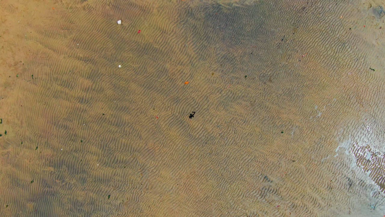 Overhead view of rippled sand in San Carlos, Baja California Sur, with small colored markers spread across the beach, a pale wet patch at lower right, and no visible people.