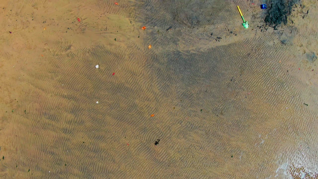 The aerial view over San Carlos sand includes rippled beach texture, a darker stained area, bright green and yellow marker-like objects near the upper right, and no visible people.
