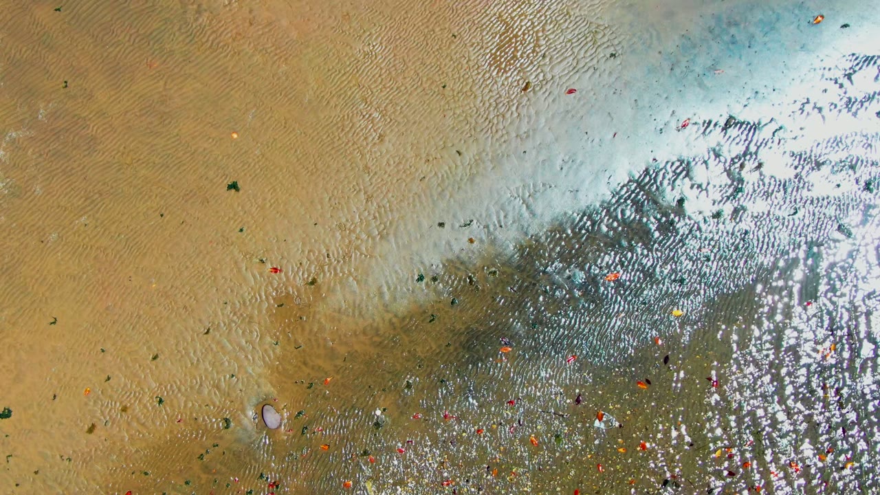 The San Carlos beach appears from above with receding shallow water on the right, exposed rippled sand on the left, small colored debris, and no visible people.