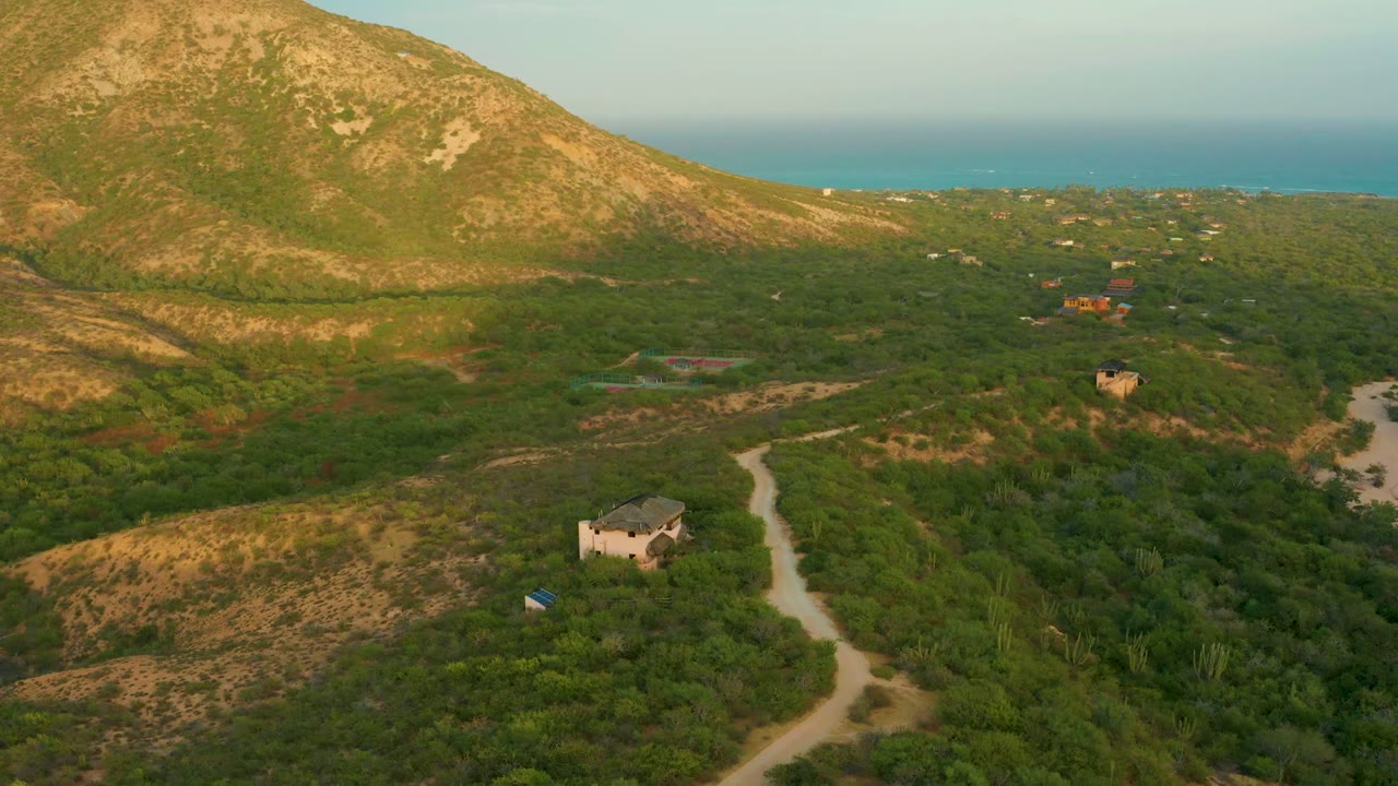 Opening aerial frame over Cabo Pulmo scrubland with a winding dirt road, a small light building near a blue tarp, tan mountain slope at left, ocean on the horizon, and no visible people.
