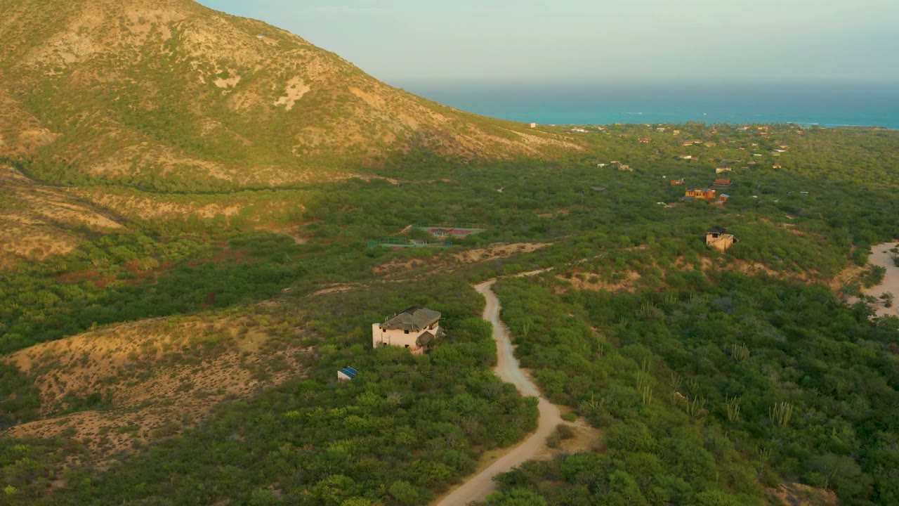 Wide aerial landscape with winding road, hillside, brush, small building, and ocean horizon.