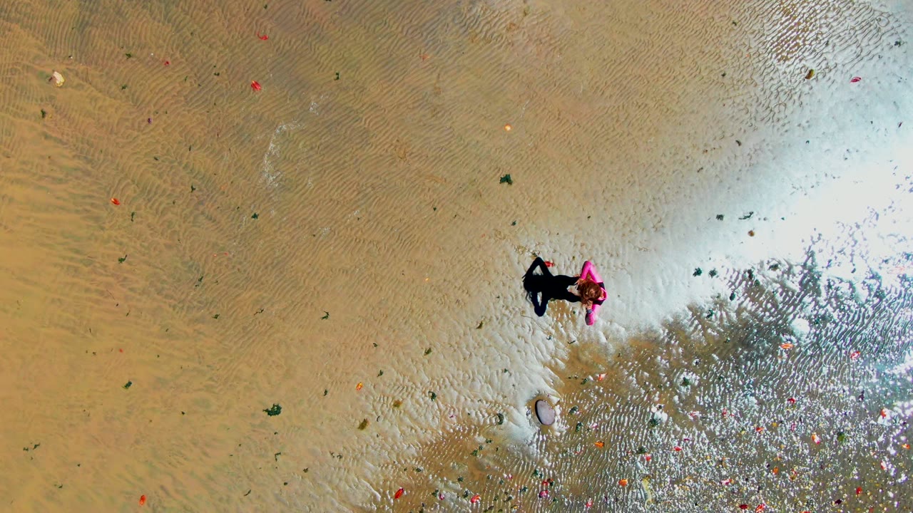 Overhead frame from San Carlos, Baja California Sur showing a person near the center-right wading through shallow rippled water, with tan sand below and small colorful debris scattered across the surface.