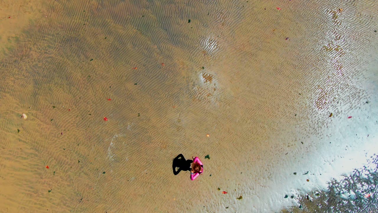 A person in pink appears right of center in the San Carlos shallow water, with a compact dark shadow, tan sand ripples, and pale reflective water along the lower right.