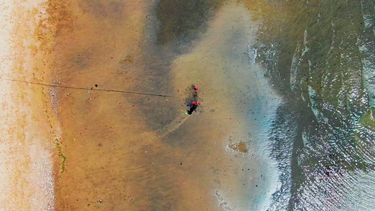 Overhead frame from San Carlos, Baja California Sur showing wet tan sand, dark damp patches, a rope or line across the beach, shallow surf at right, and small people near the center.