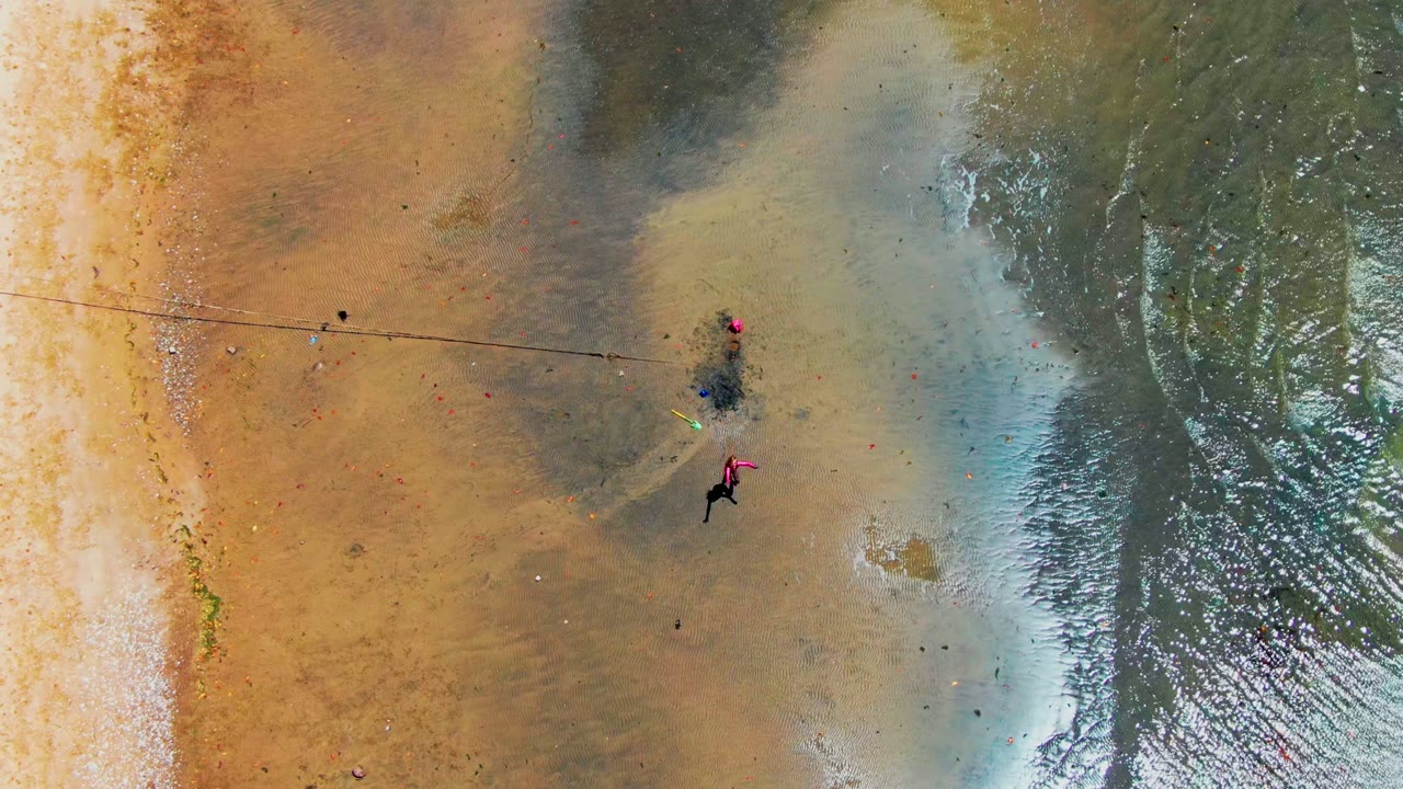 Aerial shoreline view with a person in pink moving across wet sand near shallow water.