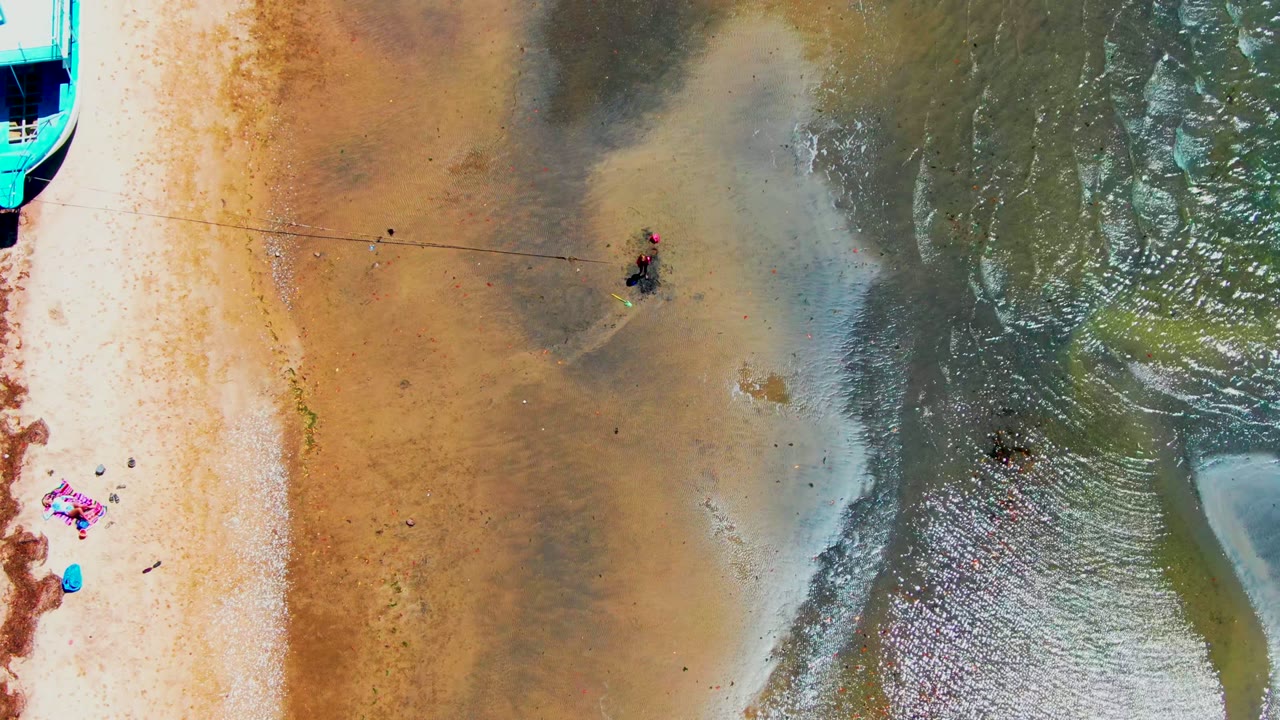 Top-down view in San Carlos, Baja California Sur, showing tan sand, green-brown shallow water, small wading figures, a colorful towel, a blue object, and a cropped turquoise boat edge.