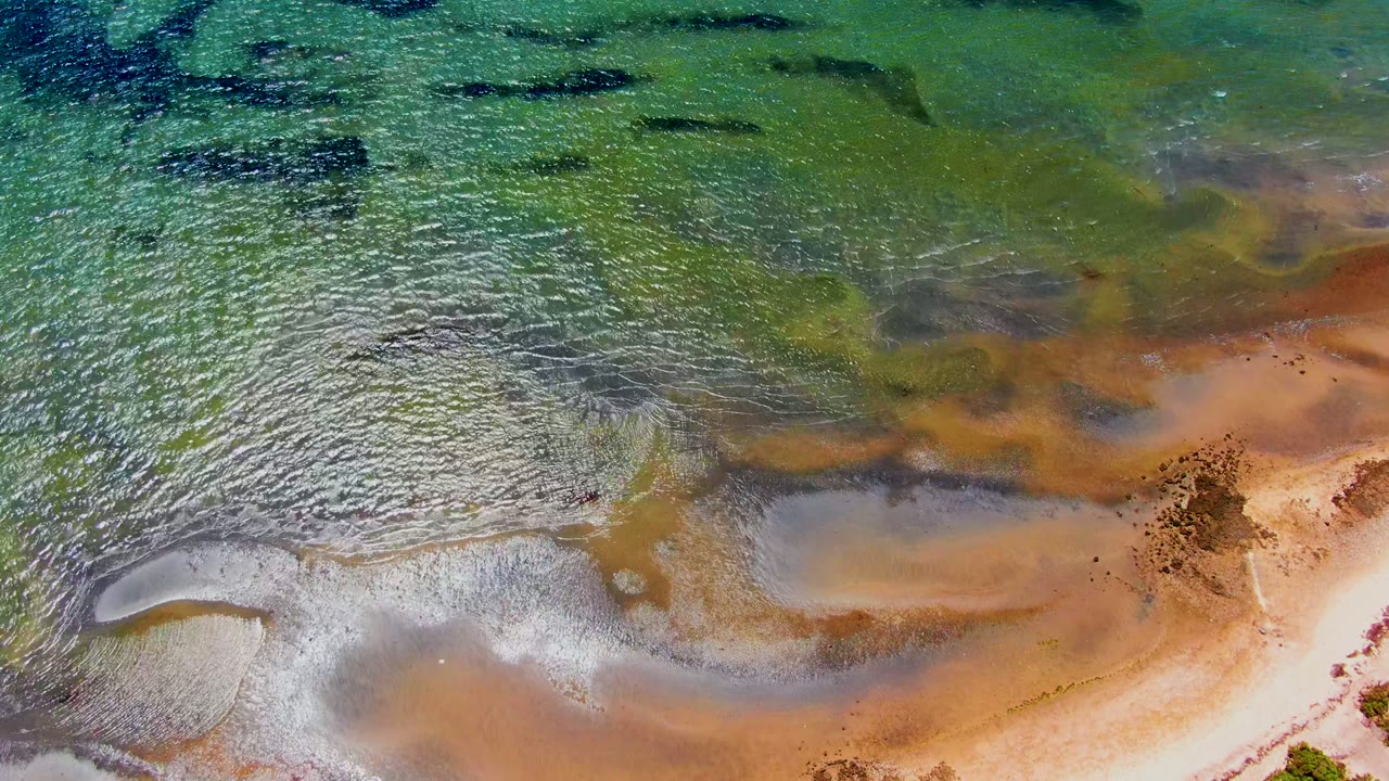 Aerial view of green-blue shallow water crossing wet sand and rust-brown tidal patterns.