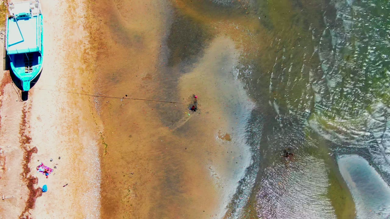 Overhead frame from San Carlos, Baja California Sur showing a bright blue boat at the sandy shore, towels lower left, and small people wading in shallow water near the center.