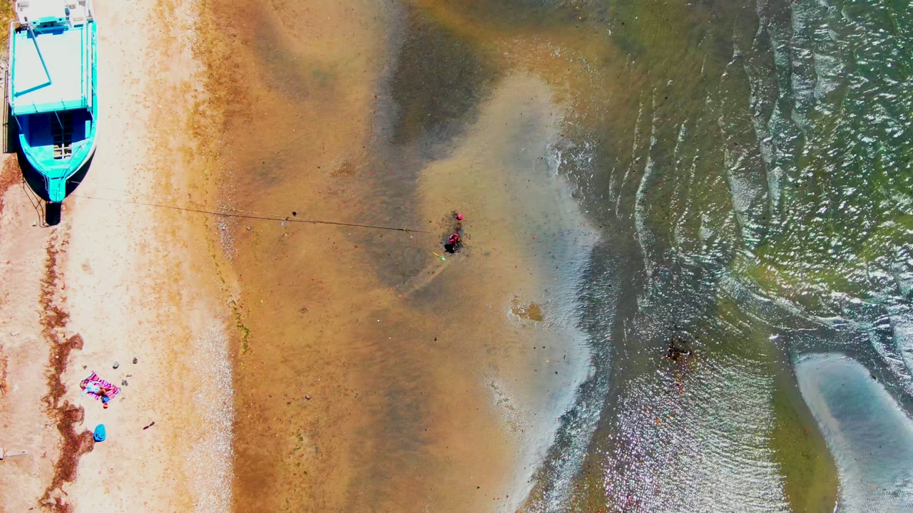 Overhead San Carlos shoreline frame showing the blue boat cropped near the top left, pale sand along the shore, and small people wading across darker wet sand and shallow water.