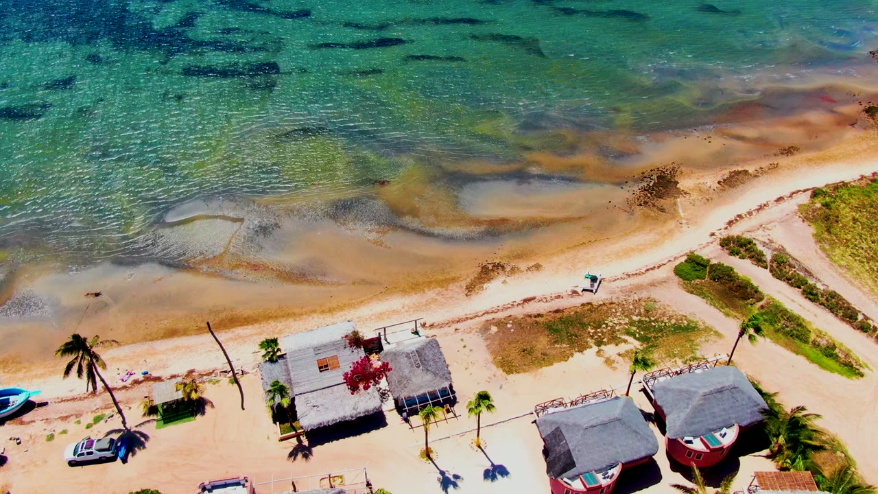 San Carlos aerial frame showing the drone farther along the shoreline, with textured shallow water above, the sand road crossing the beach, clustered huts and red buildings below, and no visible people.