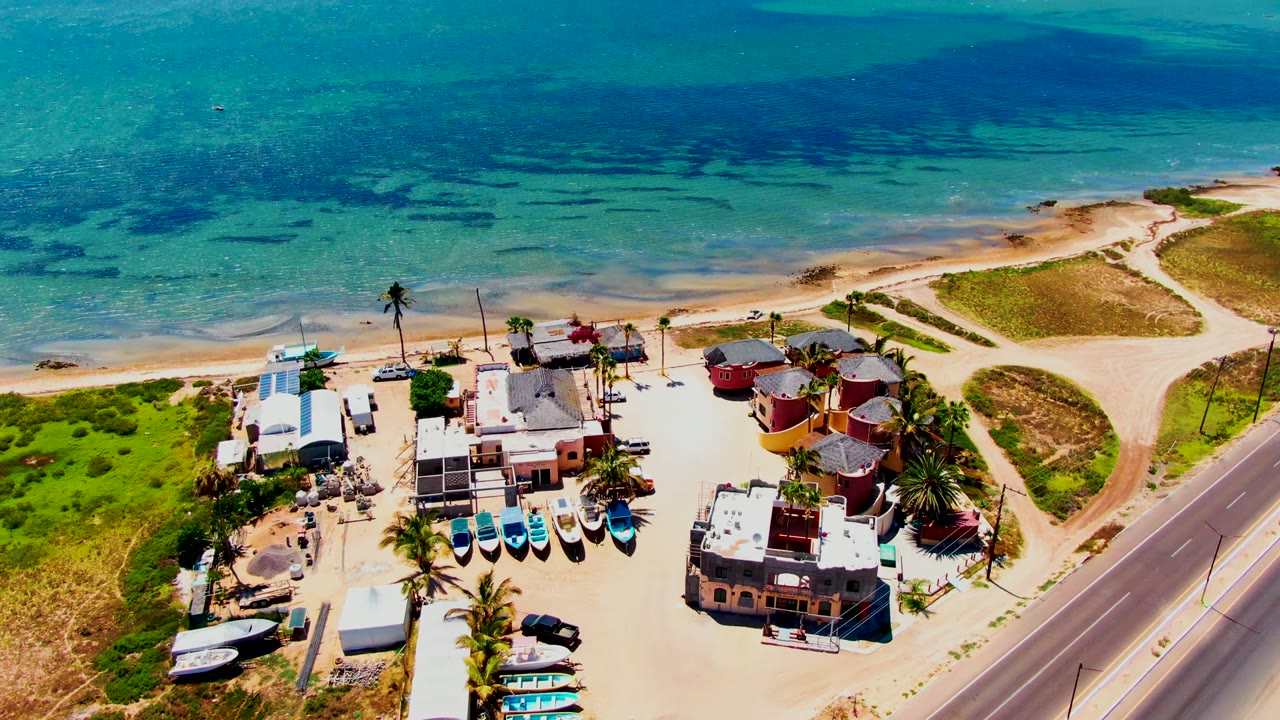 Aerial frame of San Carlos, Baja California Sur with turquoise shallow water, sandy shore, beachside buildings, parked boats, palms, a paved road, and no visible people.