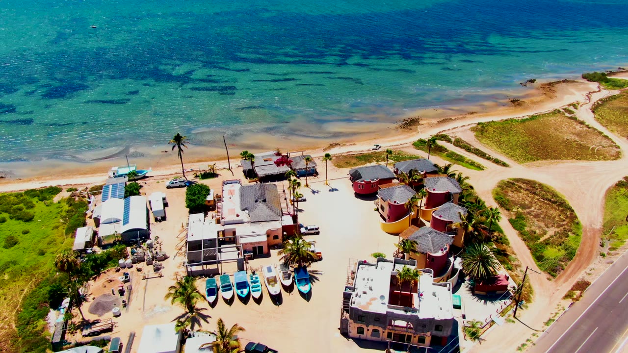 Aerial view in San Carlos with colorful round and rectangular beachside buildings, several boats, a roadside strip, sandy tracks, turquoise water, and no visible people.