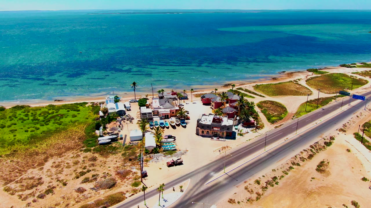 Aerial frame over San Carlos, Baja California Sur showing a coastal road, sandy beach, low buildings, parked boats, green dunes, turquoise ocean, and no visible people.